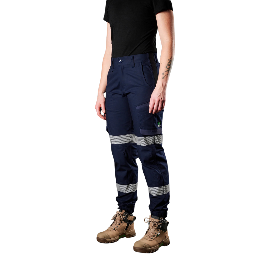 A woman standing wearing navy blue and black reflective cuffed stretch ripstop work pants with an extended back waistband and visible reflective taping.